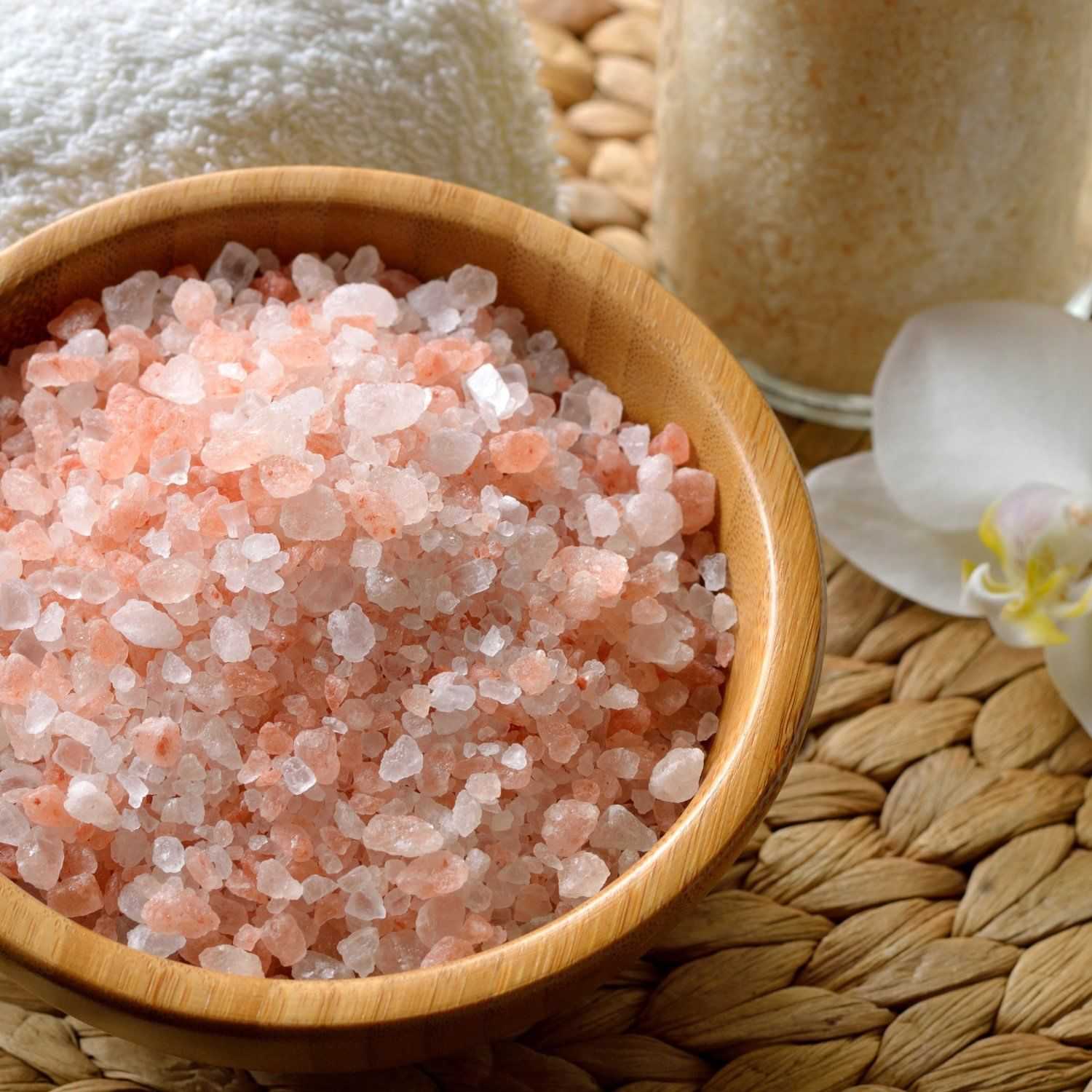Himalayan pink salt in a wooden bowl on a woven mat, near a white towel and a flower petal.