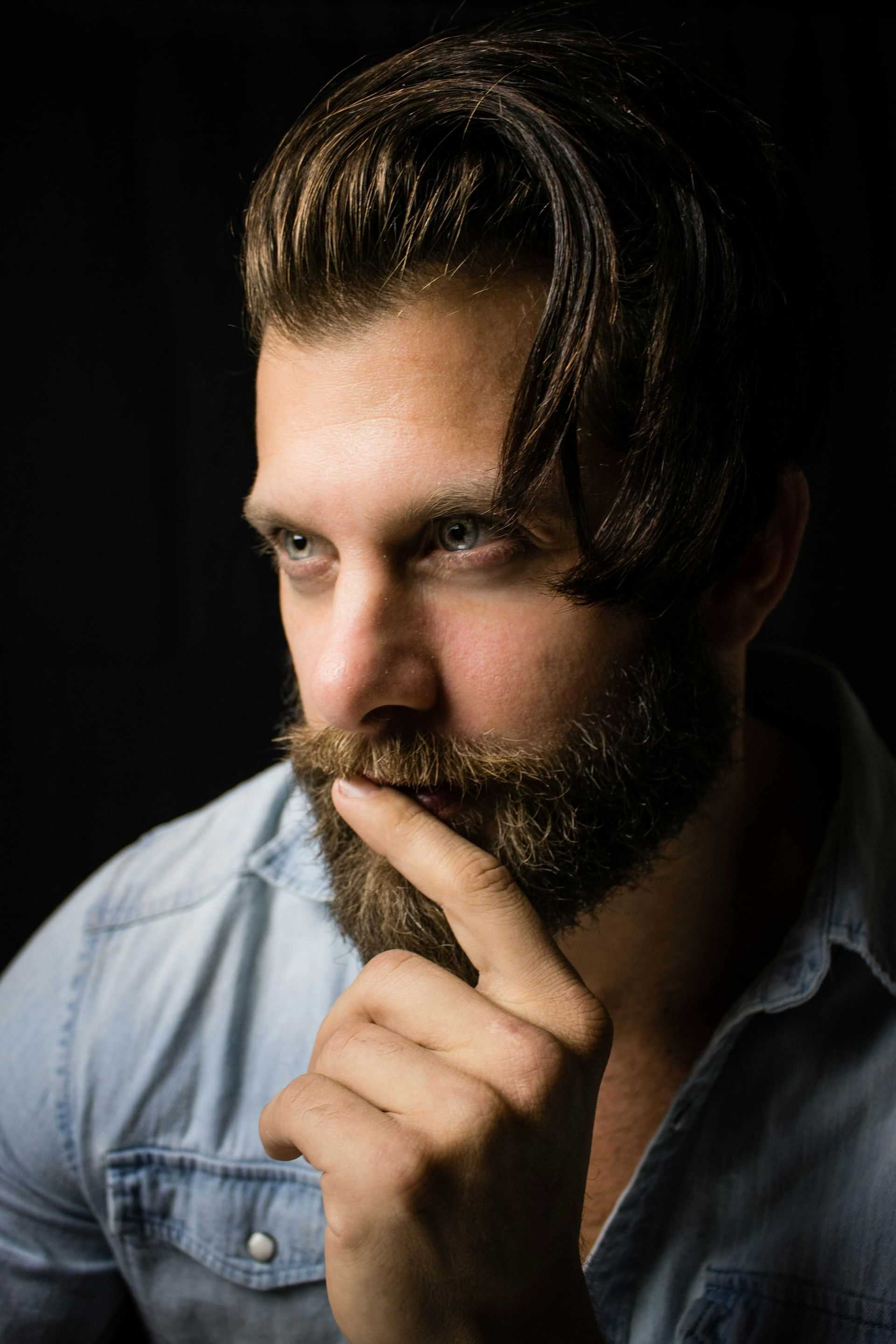 Man with a beard and thoughtful expression, wearing a denim shirt against a dark background.