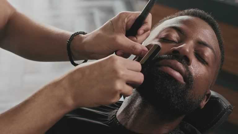 Barber carefully shaving a bearded man’s face in a barbershop.