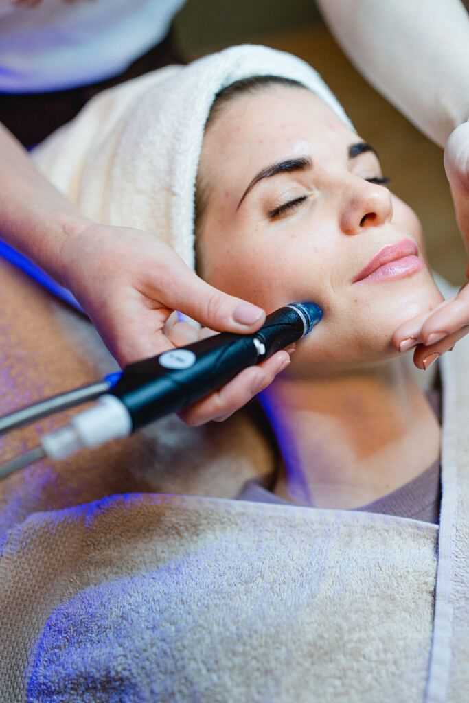 Woman enjoys a facial treatment at a spa, with a technician using a specialized skincare device.