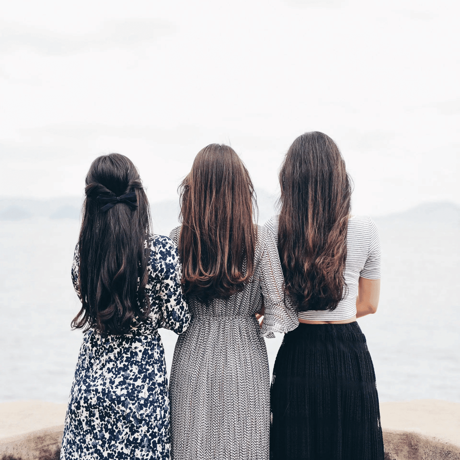 Three women with long hair standing by the sea, facing away.