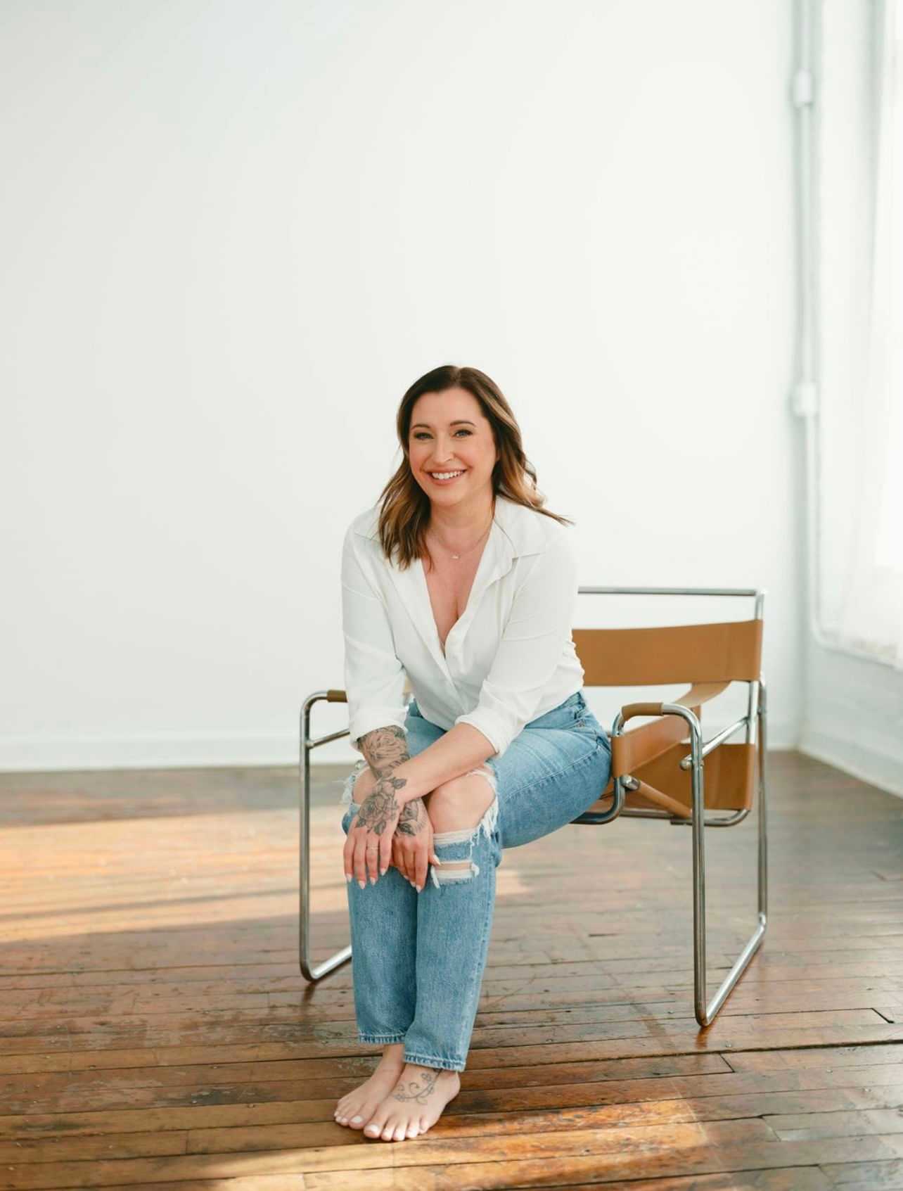 Woman sitting barefoot on chair in bright room, smiling and casually dressed in jeans and a shirt.