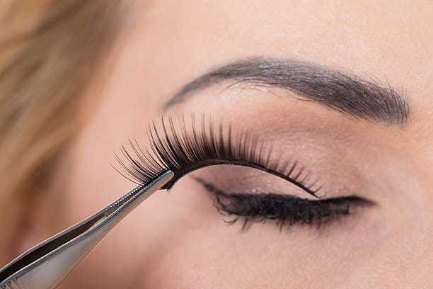 False eyelashes being placed on a closed eye with tweezers, showing precise application.