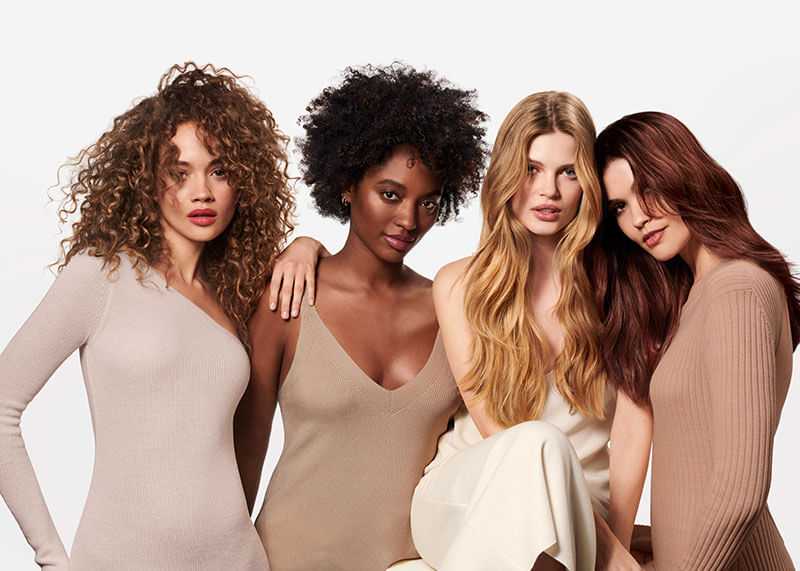 Four women with diverse hair and skin tones pose together in neutral outfits.
