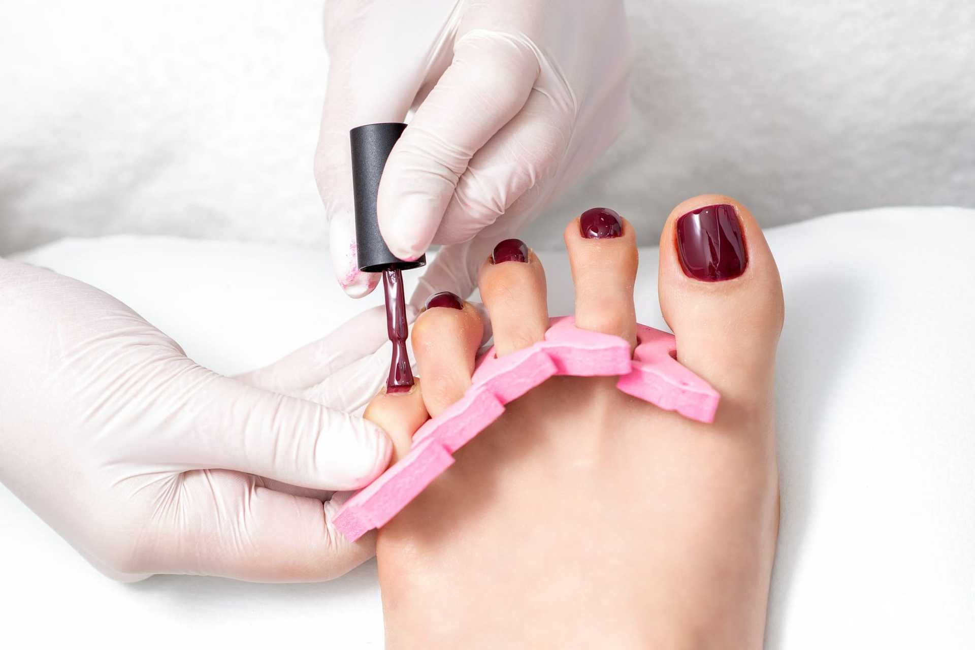Manicurist applying dark red nail polish on toes with pink toe separators in a spa setting.