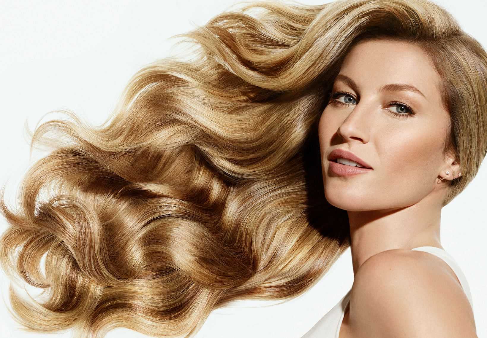 Woman with long, wavy blonde hair wearing a white top, posing against a plain background.