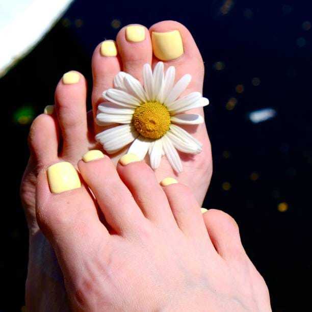 Feet with yellow nail polish and a daisy between toes, against a dark background.